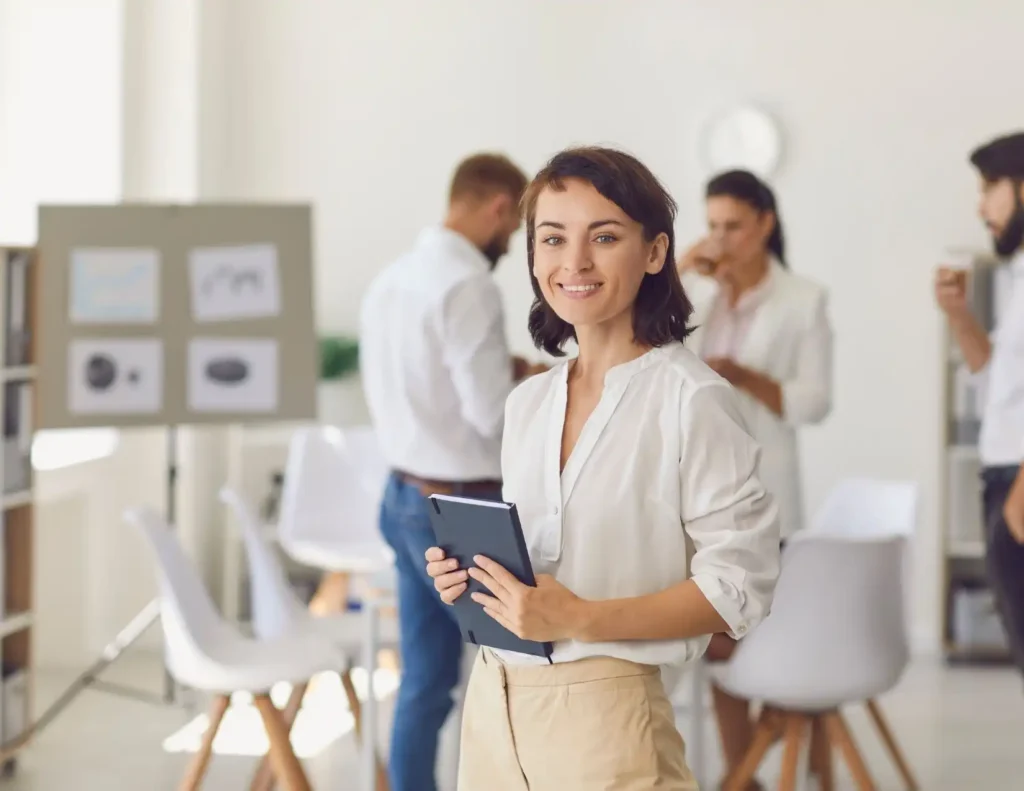 A woman dressed in professional attire smiles while standing confidently, with a group of colleagues working in the background in an office.