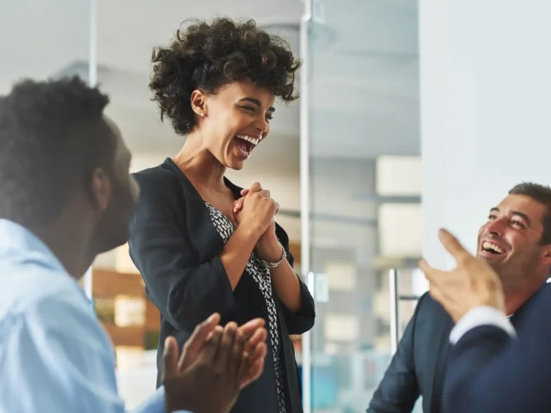 A woman joyfully claps and laughs with her colleagues, celebrating a moment of camaraderie in a professional setting.