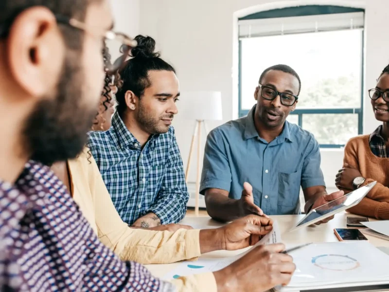 A diverse group of individuals engaged in discussion around a table, with a tablet open in front of them.