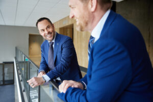 Two professional leaders in blue suits smiling and conversing while standing in a modern office hallway, symbolizing collaboration, coaching, and leadership.